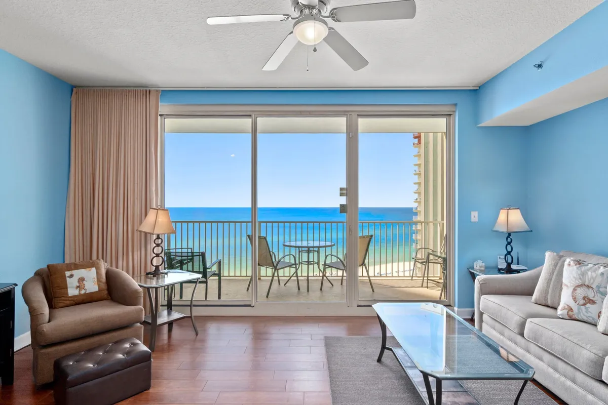 A serene master bedroom with a king-size bed adorned with decorative pillows, a large sliding glass door opening to a balcony with ocean views, and warm bedside lamps providing ambient lighting. An armchair offers a cozy reading nook, and the tranquil atmosphere is enhanced by the calming color palette and beachy decor.