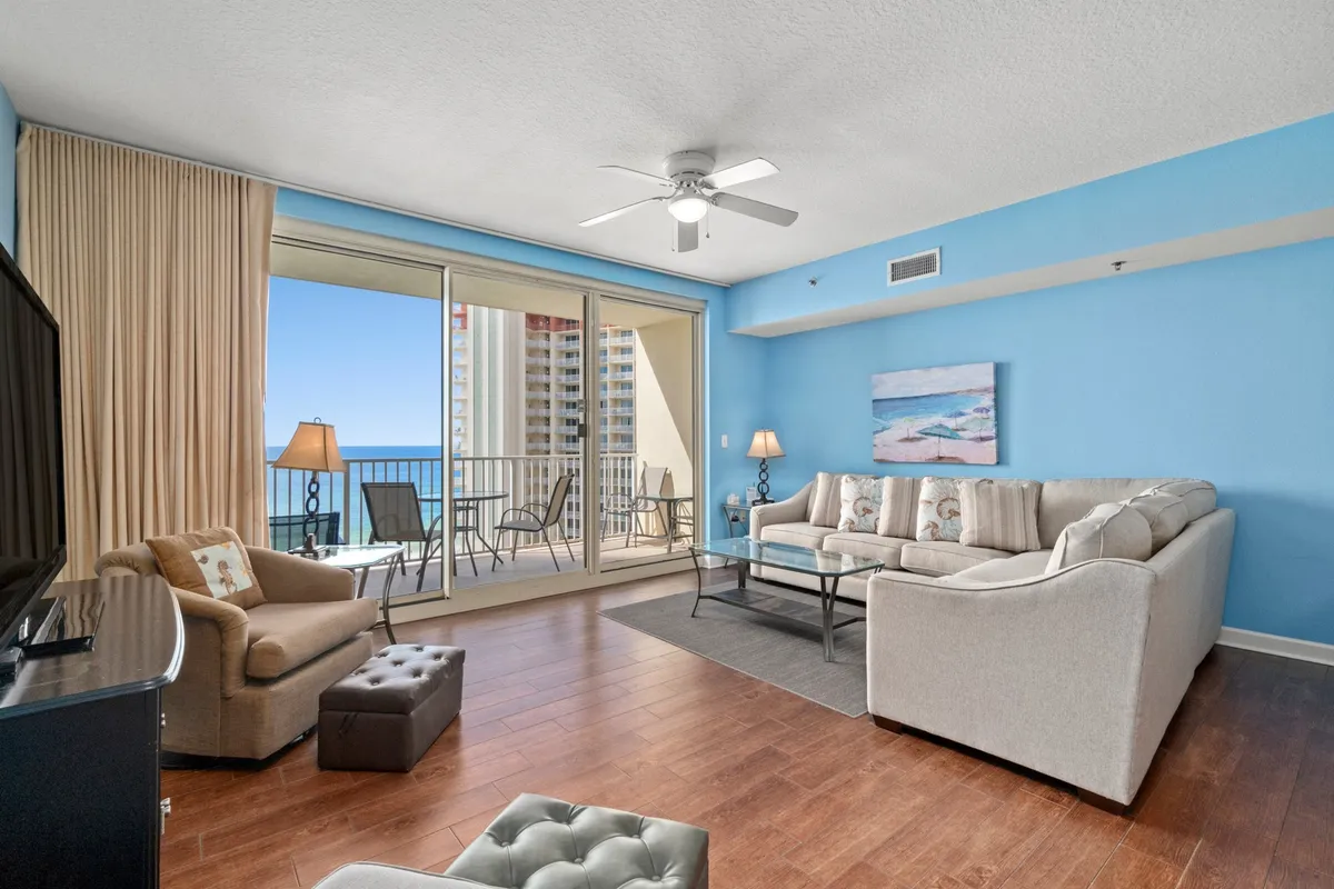 This image showcases a spacious living area featuring a comfortable sectional sofa, a flat screen television, and a large sliding glass door leading out to a balcony with ocean views. The decor is casual and coastal, with light colors and beach-themed wall art, inviting relaxation and social connection.