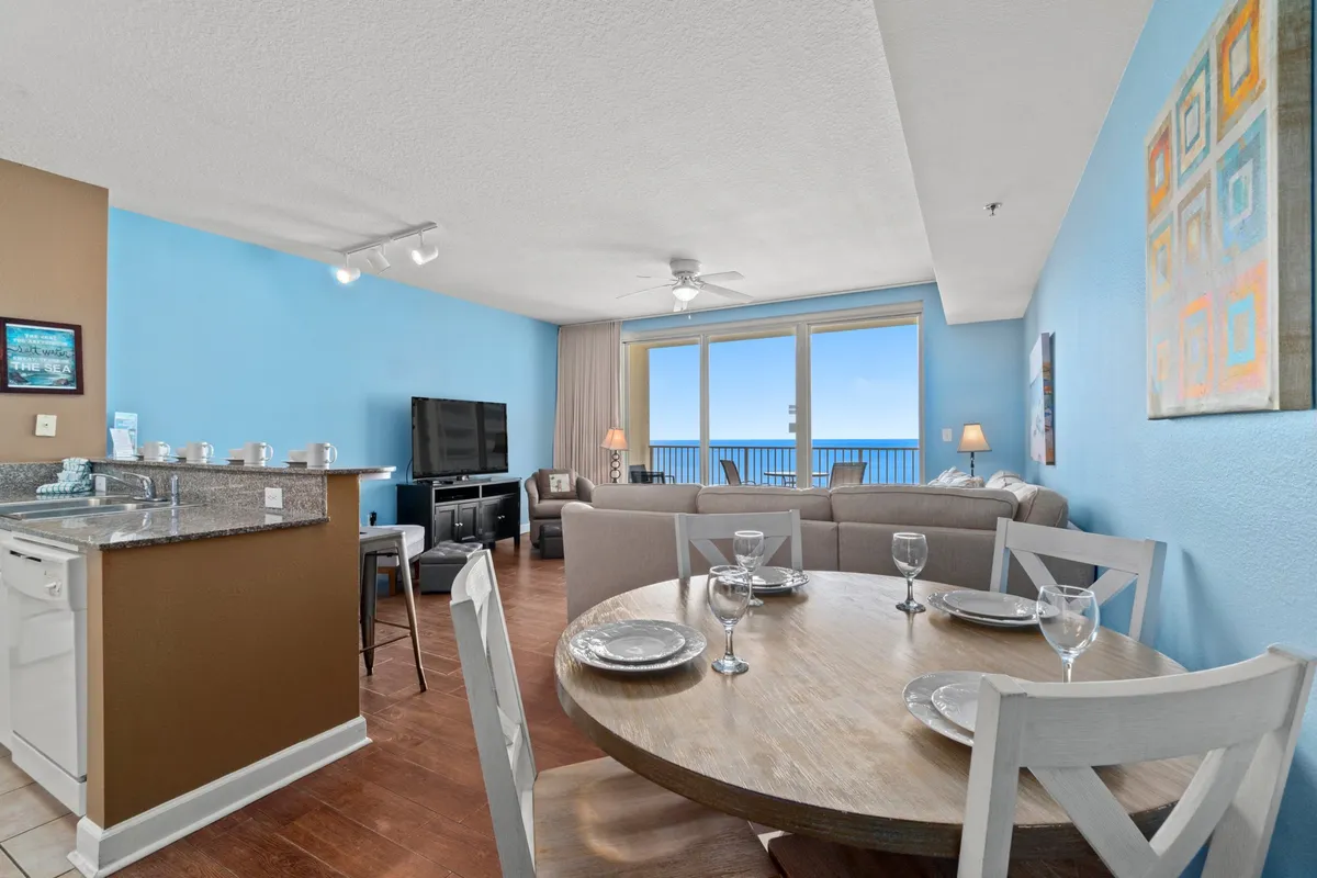 This image features a round dining table set for four, floor-to-ceiling windows offering expansive ocean views, and a modern kitchen area. The space is bathed in natural light, with light blue walls and casual, inviting decor.