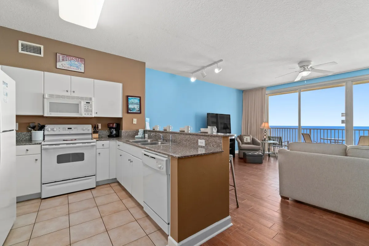 This interior image features a modern kitchen with a granite counter and bar stools, seamlessly connected to a comfortable living area with a sofa and armchair. Floor-to-ceiling windows reveal a stunning ocean view, and the kitchen is equipped with updated appliances.