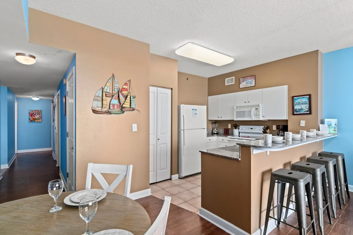 This cozy interior scene centers around a kitchen island with bar stools and a dining table set for two, set against a backdrop of coastal-themed decor. The layout invites casual dining and meal preparation, and a blue hallway leads toward other rooms, hinting at open, social spaces.