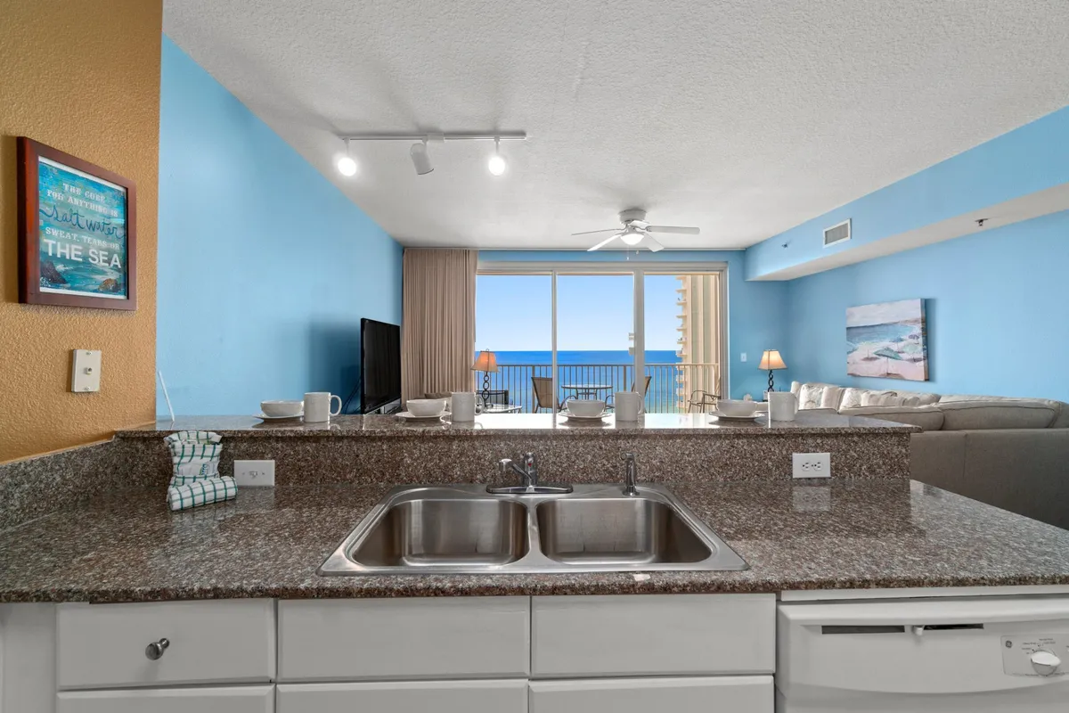 The image presents a fresh, tranquil living area with a granite kitchen countertop, double sink, and a spacious, comfortable sectional. Large sliding glass doors open to a balcony with a captivating ocean view, and a light blue accent wall enhances the coastal vibe.