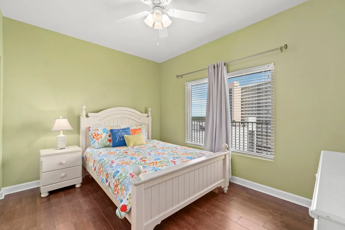 A bright, inviting bedroom featuring a white wooden bed with cheerful coral-themed bedding, a large window offering a city and resort view, and a white bedside table with a lamp for reading or relaxing. The room is filled with natural light, creating a refreshing and airy feel.