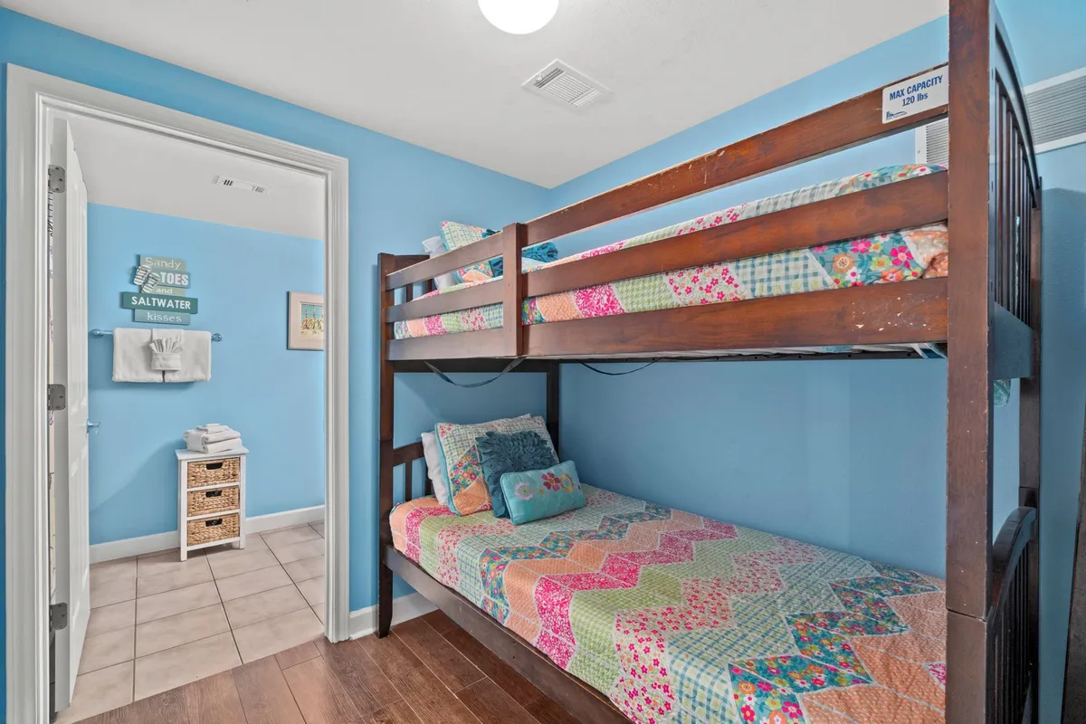 A cozy bunk area with sturdy wooden bunk beds dressed in colorful patchwork quilts. The blue-painted walls add a playful coastal touch, and an open doorway reveals an adjacent bathroom with beach-themed decor and towels, signaling convenience for younger guests.