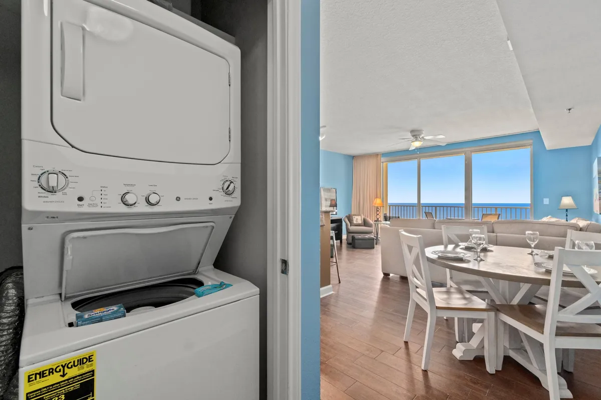 A dining table is set for a meal near floor-to-ceiling windows that open to spectacular ocean views. The image also shows a stacked washer/dryer for convenience and a living area with coastal decor, reinforcing the sense of comfort and elegance.