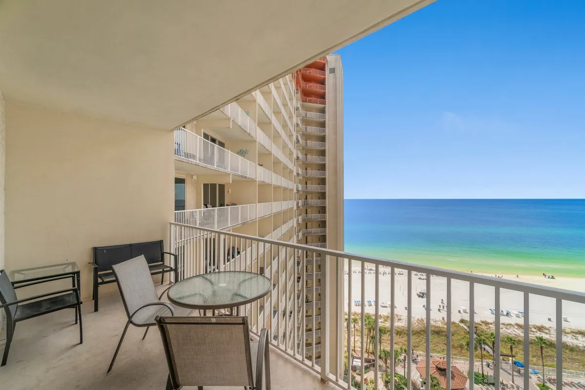 This image showcases a private balcony with a glass table and chairs, offering a sweeping, unobstructed view of the beach, ocean, and adjacent resort building. The scene is sunlit and tranquil, inviting guests to relax or dine outdoors while taking in the coastal scenery.