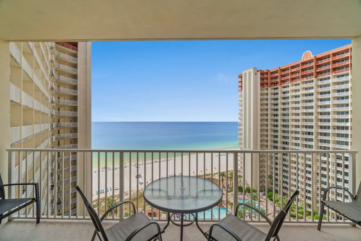 The image shows a private balcony with a glass-top table and chairs, overlooking the beach, ocean, and resort pool area. The balcony invites relaxation and socializing, with the ocean horizon providing a stunning backdrop.