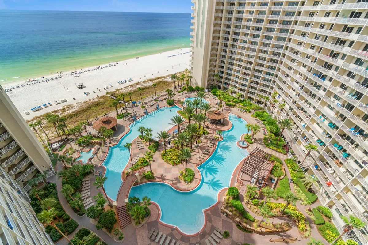 A panoramic view shows the lagoon-style pool with fountains, the tall resort building, private beach access with white sand, and lush palm trees and gardens. The scene captures guests swimming, lounging, and enjoying both the pool and the adjacent private beach area.