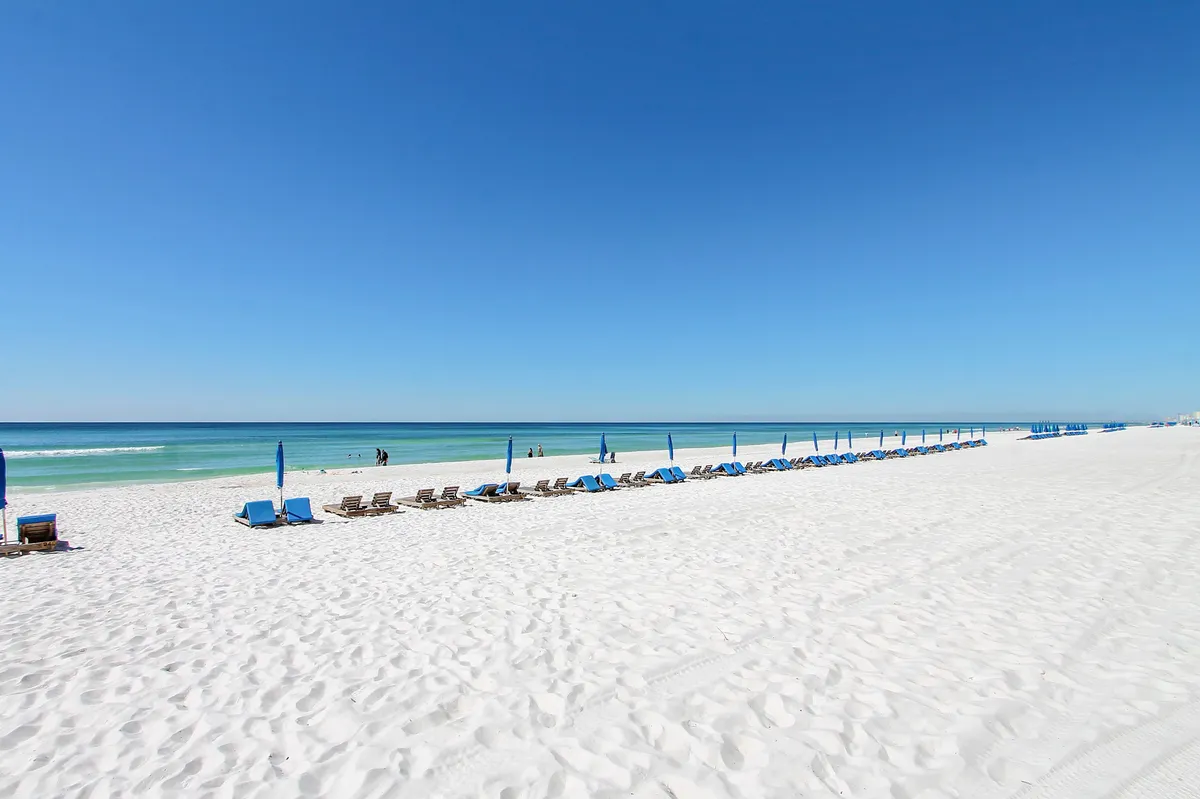 A picturesque line of blue beach umbrellas and lounge chairs is set along a pristine white sandy beach. The calm, turquoise ocean stretches out to the horizon beneath a clear blue sky, inviting guests to relax, swim, or stroll along the shore.