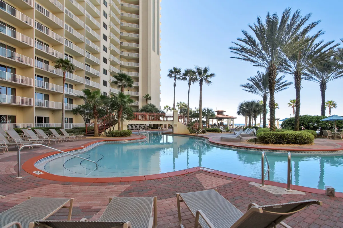 A large, lagoon-style swimming pool framed by tall palm trees and high-rise resort balconies. Lounge chairs and umbrellas line the brick pool deck, and the inviting water is the centerpiece. The atmosphere reflects leisure, relaxation, and tropical resort luxury.