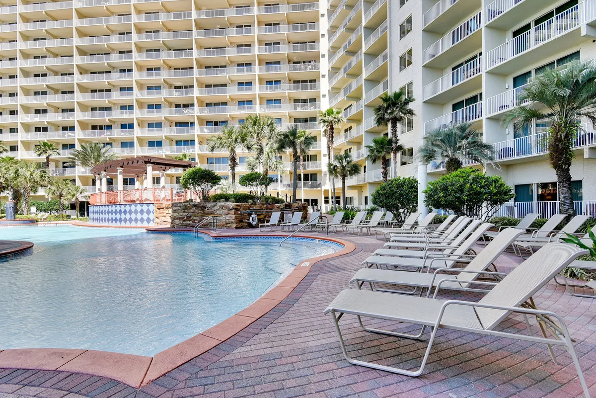 The photo captures a vibrant lagoon-style swimming pool with fountains, a row of lounge chairs, a tiki bar structure, and a tall resort building with balconies. The atmosphere is sunny and leisurely, emphasizing both relaxation and social opportunities at the tiki bar.