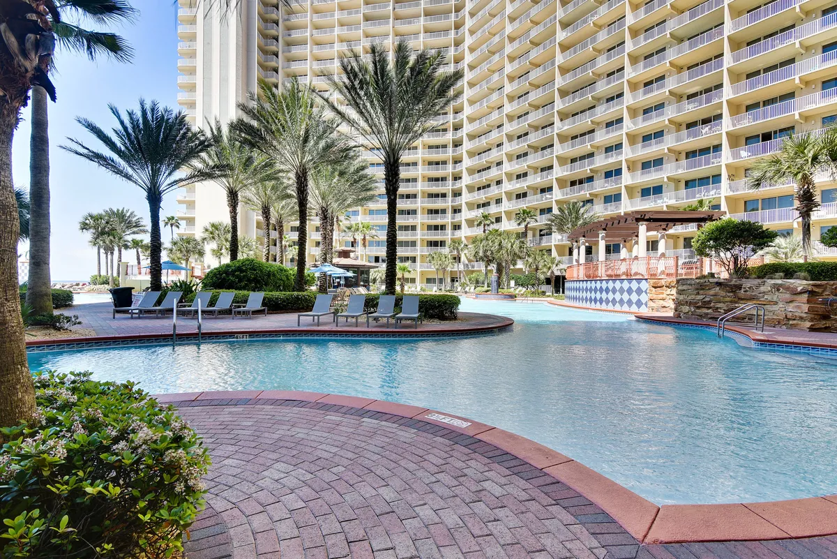 A stunning lagoon-style resort pool is surrounded by tall palm trees and bordered by a modern high-rise building with balconies. There are poolside lounge chairs and shaded seating areas, creating a luxurious and inviting poolside atmosphere. The scene is lively yet peaceful, with clear blue water and ample space for relaxation and socializing.