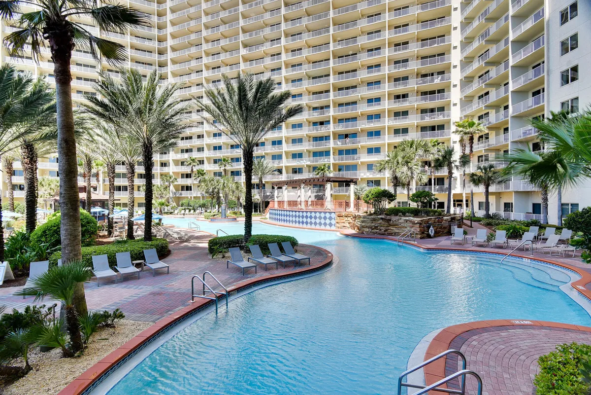 A sweeping view of the Shores of Panama's signature lagoon-style swimming pool, complete with playful fountains, palm trees, and a resort building with balconies overlooking the pool. Lounge chairs are arranged along the poolside, inviting families to relax and soak up the sun.