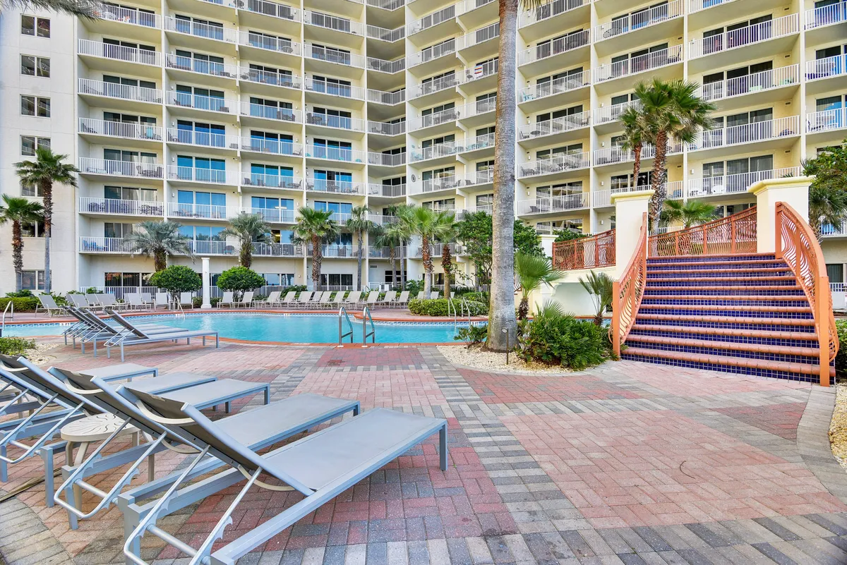 A wide, lagoon-style swimming pool sits at the heart of the resort, surrounded by rows of lounge chairs, tall palm trees, and ornate tiled staircases. The setting is sun-drenched and tropical, with space for groups to relax and socialize poolside.