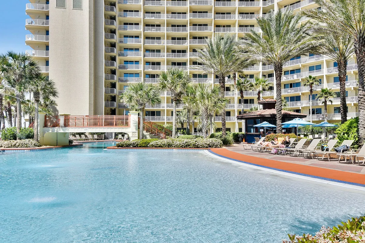 A stunning lagoon-style swimming pool sits at the heart of a resort, surrounded by lush palm trees and fringed with lounge chairs and umbrellas. Balconies from the high-rise condo overlook the pool area, giving a sense of the resort's amenities and luxury.