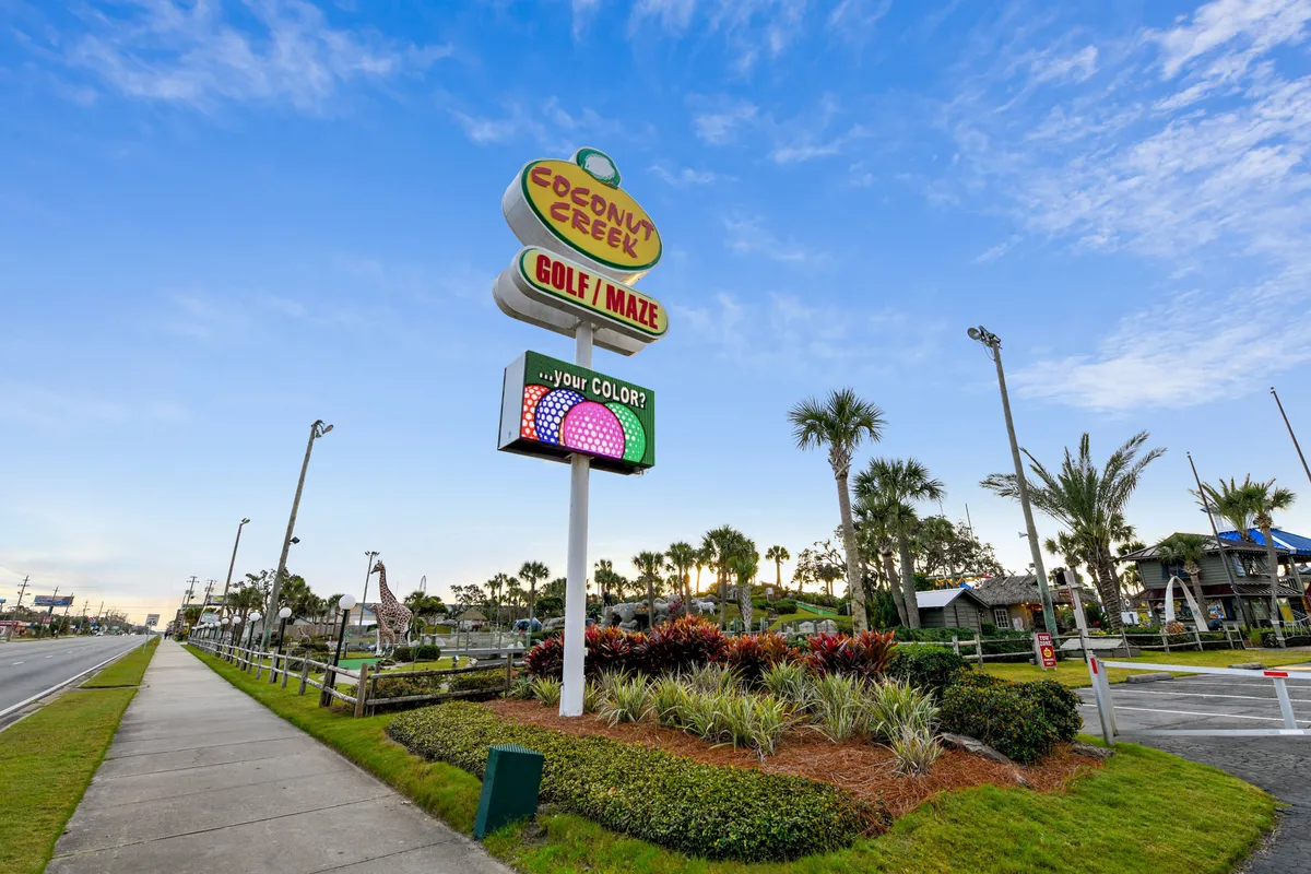 A colorful and lively miniature golf course is framed by palm trees and playful decorations. The Coconut Creek Golf / Maze sign is visible, and people are seen enjoying outdoor activities. The landscape is vibrant and family-friendly, evoking a sense of fun and seasonal recreation.