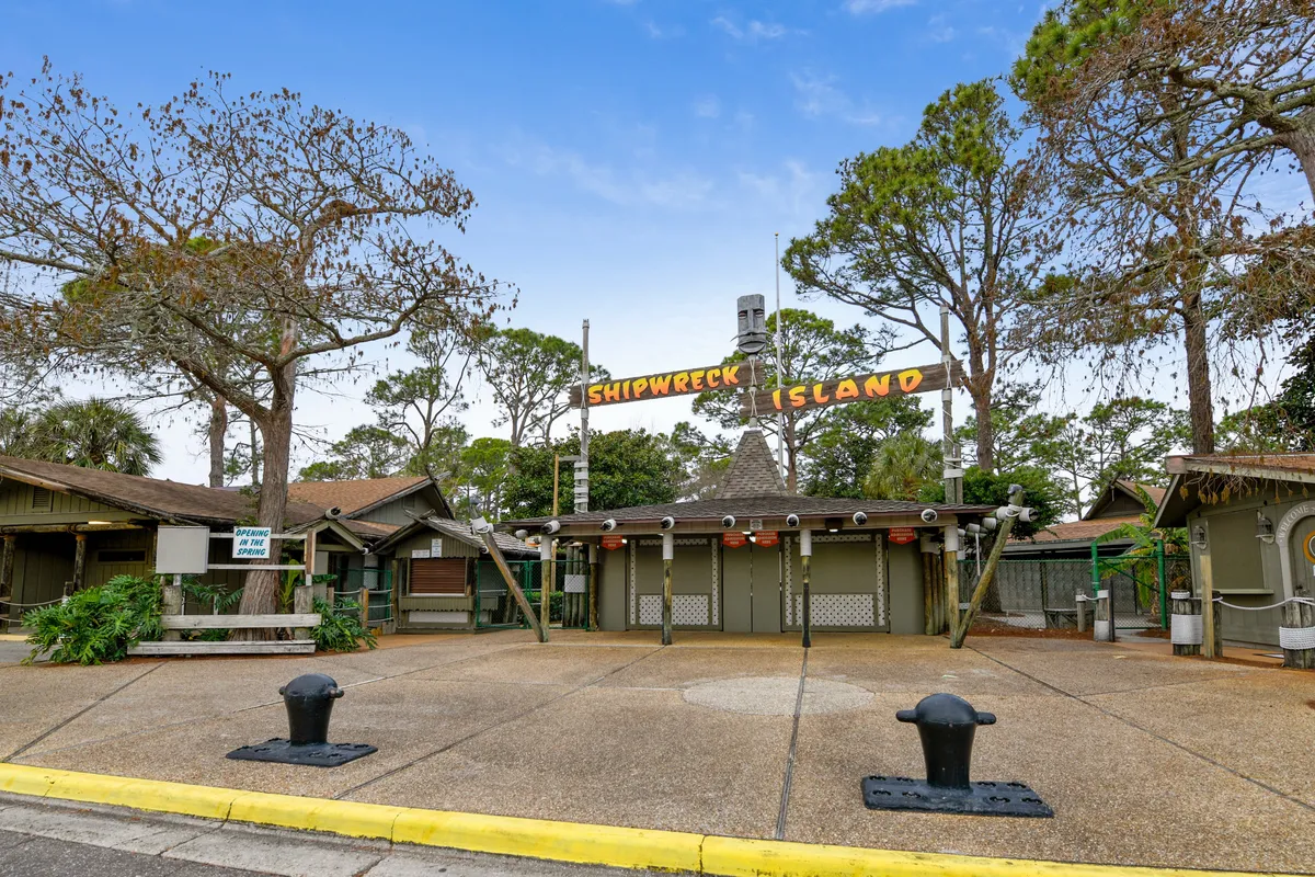 The pirate-themed entrance sign for Shipwreck Island water park, surrounded by rustic wooden buildings and mature trees. The area buzzes with excitement and anticipation as families prepare for a day of waterslides and aquatic adventures.